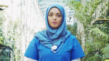 Professional Muslim medical doctor works in front of hospital office. Portrait of young and attractive Middle Eastern female physician in hijab. Medical concept.