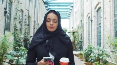 Portrait of young and confident Muslim business woman with a smartphone and coffee in hijab. Middle Eastern office worker or businesswoman is working outdoor in the city. Corporate and financial