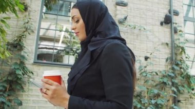 Portrait of young and confident Muslim business woman with a smartphone and coffee in hijab. Middle Eastern office worker or businesswoman is working outdoor in the city. Corporate and financial