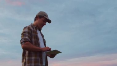Farmer with a tablet computer in front of a sunset agricultural landscape. Man in a countryside field. The concept of country life, food production, farming and technology.
