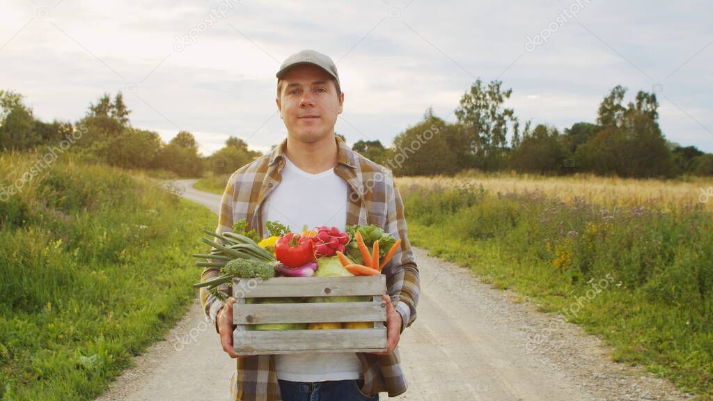 Farmer with a vegetable box in front of a sunset agricultural landscape. Man in a countryside field. The concept of country life, food production, farming and country lifestyle.