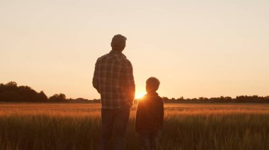 Farmer and his son in front of a sunset agricultural landscape. Man and a boy in a countryside field. The concept of fatherhood, country life, farming and country lifestyle.