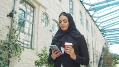 Portrait of young and confident Muslim business woman with a smartphone and coffee in hijab. Middle Eastern office worker or businesswoman is working outdoor in the city. Corporate and financial