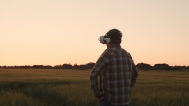 Farmer in virtual reality helmet in front of a sunset agricultural landscape. Man in a countryside field. The concept of country life, food production, farming and technology.