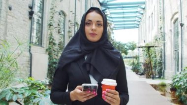 Portrait of young and confident Muslim business woman with a smartphone and coffee in hijab. Middle Eastern office worker or businesswoman is working outdoor in the city. Corporate and financial