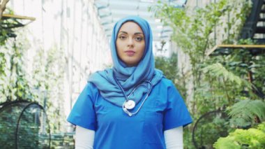 Professional Muslim medical doctor works in front of hospital office. Portrait of young and attractive Middle Eastern female physician in hijab. Medical concept.