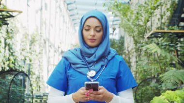 Professional Muslim medical doctor works in front of hospital office. Portrait of young and attractive Middle Eastern female physician in hijab. Medical concept.