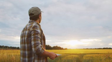 Farmer with a tablet computer in front of a sunset agricultural landscape. Man in a countryside field. The concept of country life, food production, farming and technology.