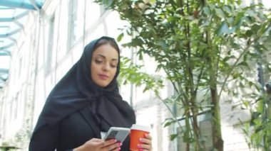 Portrait of young and confident Muslim business woman with a smartphone and coffee in hijab. Middle Eastern office worker or businesswoman is working outdoor in the city. Corporate and financial