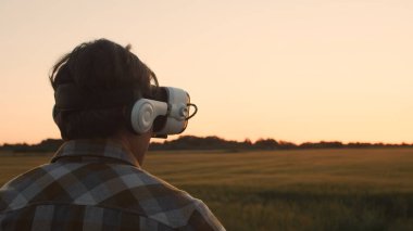 Farmer in virtual reality helmet in front of a sunset agricultural landscape. Man in a countryside field. The concept of country life, food production, farming and technology.