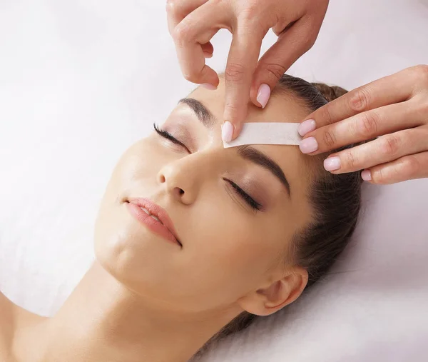 Professional worker is removing hair from young and beautiful female face with hot wax. Girl has a beauty treatment procedure. Depilation, epilation, skin and health care concepts.
