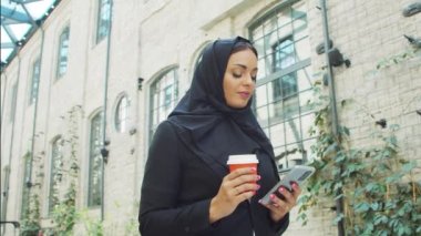 Portrait of young and confident Muslim business woman with a smartphone and coffee in hijab. Middle Eastern office worker or businesswoman is working outdoor in the city. Corporate and financial