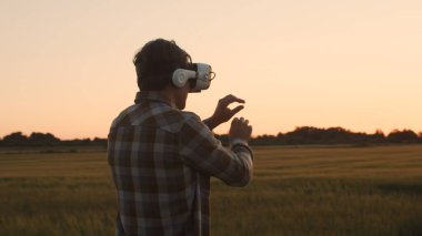 Sanal gerçeklik kaskı takan çiftçi, gün batımı tarımının önünde. Kırsal kesimde bir adam. Kır hayatı, gıda üretimi, tarım ve teknoloji kavramı.