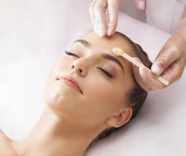 Profesyonel işçi, genç ve güzel kadın yüzünden sıcak balmumuyla saç kesiyor. Kızın güzellik tedavi prosedürü var. Defilasyon, epilasyon, cilt ve sağlık konseptleri.