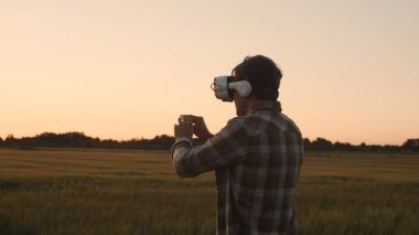 Sanal gerçeklik kaskı takan çiftçi, gün batımı tarımının önünde. Kırsal kesimde bir adam. Kır hayatı, gıda üretimi, tarım ve teknoloji kavramı.