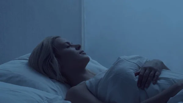 Young woman lying in the bed at night and having insomnia disease. Beautiful blond sleeping girl. Twilight in the bedroom, moonlight from the window. The concept of sleeplessness, health and rest.