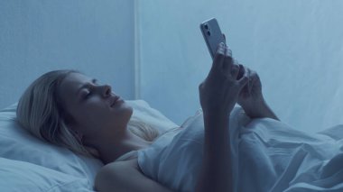 Genç bir kadın gece akıllı telefonuyla yatakta yatıyor. Güzel sarışın, dinlenen kız. Yatak odasında alacakaranlık, pencereden ay ışığı. Sağlık ve dinlenme kavramı.