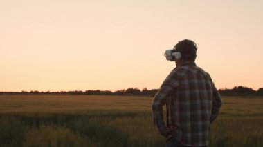 Farmer in virtual reality helmet in front of a sunset agricultural landscape. Man in a countryside field. The concept of country life, food production, farming and technology.