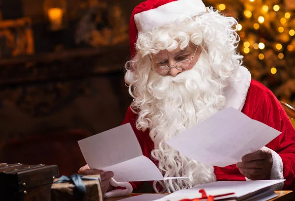 Workplace of Santa Claus. Cheerful Santa is reading letters from children while sitting at the table. Fireplace and Christmas Tree in the background. Traditional Christmas concept.