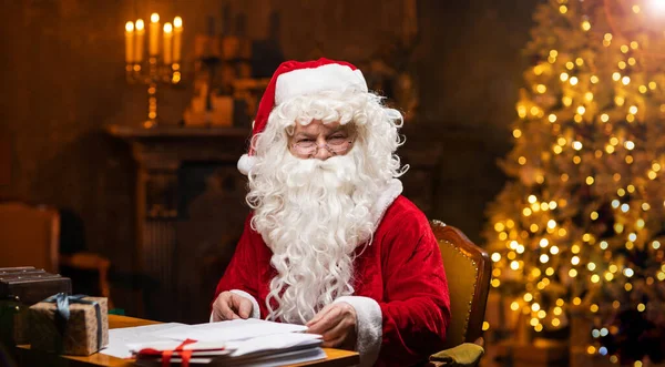 サンタクロースの職場 陽気サンタはテーブルに座って手紙を書いています 背景にある暖炉とクリスマスツリー 伝統的なクリスマスのコンセプト — ストック写真