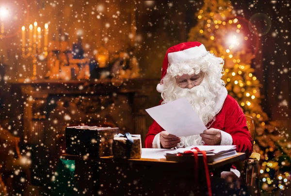 サンタクロースの職場 陽気サンタはテーブルに座っている間に子供たちからの手紙を読んでいます 背景にある暖炉とクリスマスツリー 伝統的なクリスマスのコンセプト — ストック写真