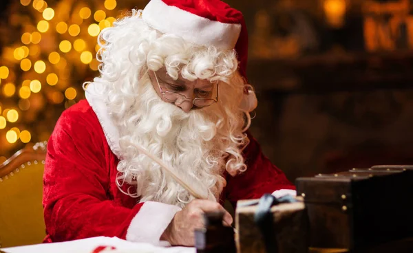 Workplace of Santa Claus. Cheerful Santa is writing the letter while sitting at the table. Fireplace and Christmas Tree in the background. Traditional Christmas concept.