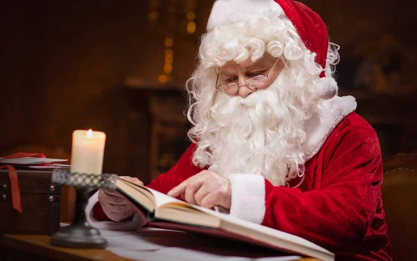 Workplace of Santa Claus. Cheerful Santa is reading the book of wishes while sitting at the table. Fireplace and Christmas Tree in the background. Traditional Christmas concept.