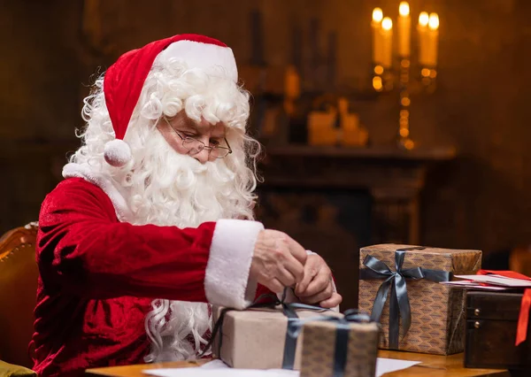 Workplace of Santa Claus. Cheerful Santa is wrapping the gift while sitting at the table. Fireplace and Christmas Tree in the background. Traditional Christmas concept.