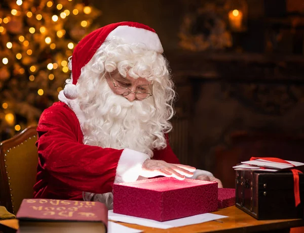 Workplace of Santa Claus. Cheerful Santa is conjuring over the gift box while sitting at the table. Fireplace and Christmas Tree in the background. Traditional Christmas concept.