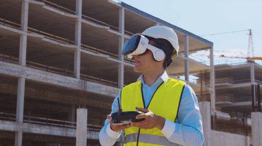 Sanal gerçeklik kasklı profesyonel dron operatörü inşaat alanının önünde duruyor. Uzaktan kumandalı bir bina. Ofis binası ve vinç arka planı. İş, gayrimenkul ve