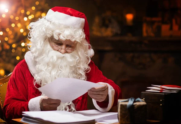 Workplace of Santa Claus. Cheerful Santa is reading letters from children while sitting at the table. Fireplace and Christmas Tree in the background. Traditional Christmas concept.