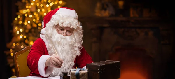 サンタクロースの職場 陽気サンタはテーブルに座って手紙を書いています 背景にある暖炉とクリスマスツリー 伝統的なクリスマスのコンセプト — ストック写真