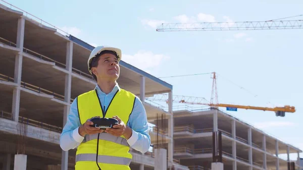 Drone operator holding remote controller. Professional builder in helmet and vest standing in front of construction site. Office building and crane background. Business, real estate and investment