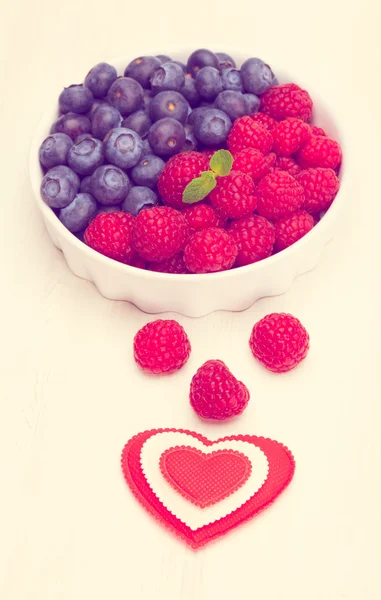 Raspberry bilberry in white bowls and heart. Toned in warm colors — Stock Photo, Image