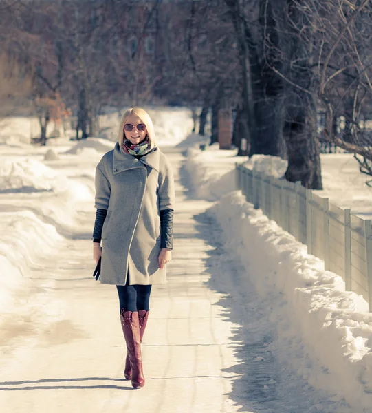 Mooie jonge blonde vrouw wandelen in het park in Winternamiddag in rode laarzen, jas en zonnebril. — Stockfoto