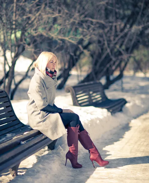 Linda jovem loira sentada em um banco de casaco de inverno e botas vermelhas. Inverno noite ensolarada . Imagem De Stock