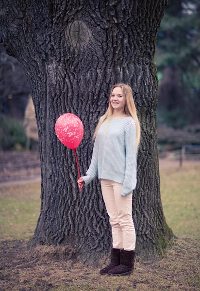 Open portrait of the young woman the beautiful woman in cold weather in park. The sensual blonde poses and is cheerful with a red balloon. — Stock Photo, Image