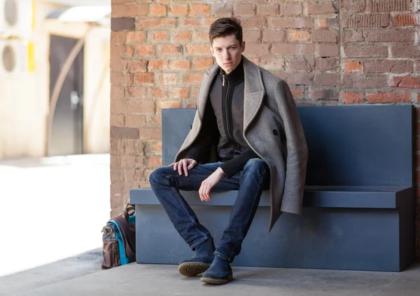 Posiert der junge Mann im Mantel auf einer Bank sitzend — Stockfoto