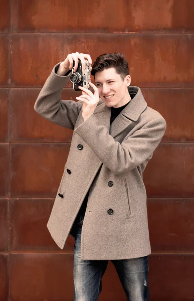 Молодой человек в пальто фотографирует макет винтажной камеры на металлической ржавой стене — стоковое фото