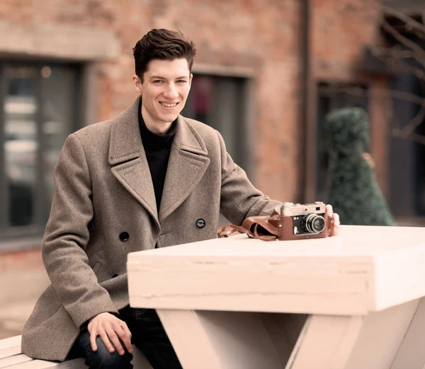 Het model van de jonge man in een vacht zit aan een witte tafel met de vintage camera buitenshuis — Stockfoto