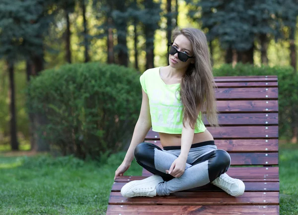 La modelo hermosa muchacha fitness se sienta en las gafas de sol sobre la silla de madera en el parque. Pantalones deportivos cortos. Abdominales fitness —  Fotos de Stock