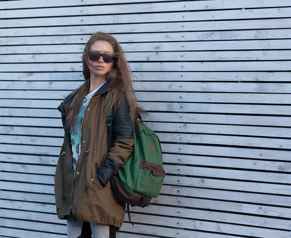 Menina bonita Cabelo longo. Casaco de turista, óculos de sol, mochila. parede de madeira branca . — Fotografia de Stock