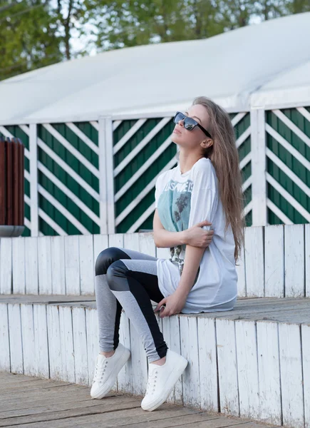 La hermosa joven con el pelo largo en gafas de sol se sienta en los pasos de madera blanca en el verano — Foto de Stock