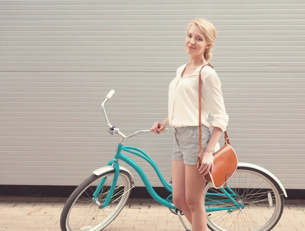 Hermosa chica rubia está de pie cerca de la bicicleta vintage con bolso vintage marrón divertirse y buen humor mirando en la cámara y sonriendo, cálido, tonificación —  Fotos de Stock