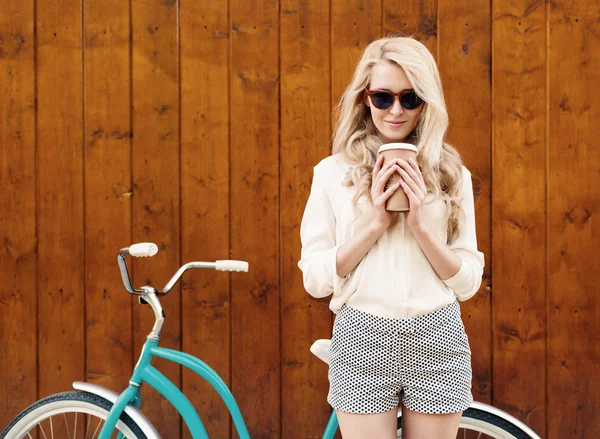 Ung sexig blond tjej med långt hår i Solglasögon står nära vintage grön cykel och hålla en kopp kaffe har kul och gott humör tittar i kameran och leende, värma, Tönning — Stockfoto