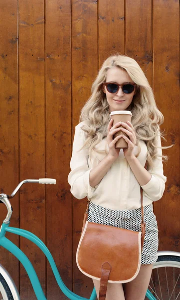 Ung sexig blondin tjej med långt hår med brun vintage väska i Solglasögon står nära vintage grön cykel och hålla en kopp kaffe, ha kul och gott humör titta i kameran och leende, varm, — Stockfoto