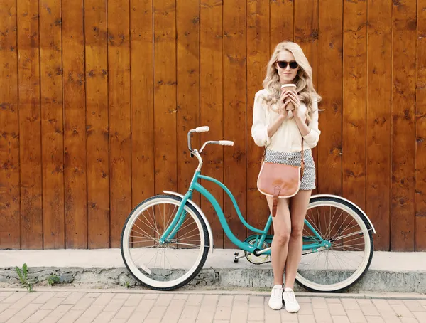 Joven chica rubia sexy con el pelo largo con bolso vintage marrón en gafas de sol de pie cerca de la bicicleta verde vintage y sosteniendo una taza de café, divertirse y buen humor mirando en la cámara y sonriente, cálido , — Foto de Stock