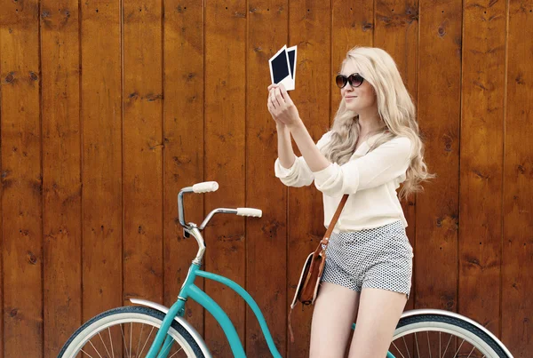 Joven mujer rubia sexy de pie cerca de una bicicleta verde vintage celebración de fotos y sonriente, cálido, tonning —  Fotos de Stock