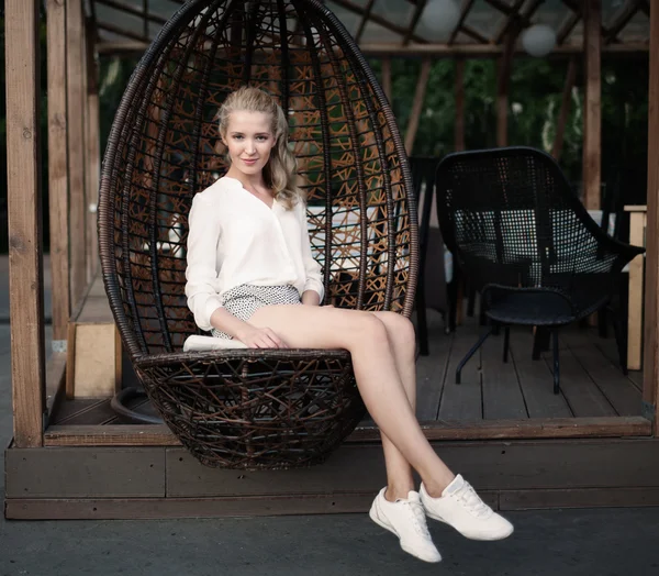 Mulher loira jovem bonita com pernas longas sentado em uma cadeira de vime em um café ao ar livre em uma noite quente de verão, sorrindo e olhando para a câmera — Fotografia de Stock