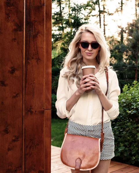 Ung sexig blondin tjej med långt hår i solglasögon med brun vintage väska håller en kopp kaffe har roligt och bra humör titta i kameran och leende, varm, Tönning — Stockfoto